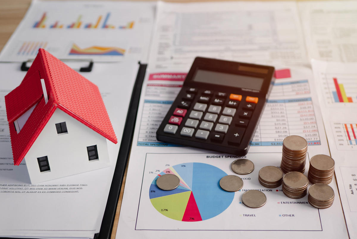 A calculator, coins and financial charts on a desk beside a small model house