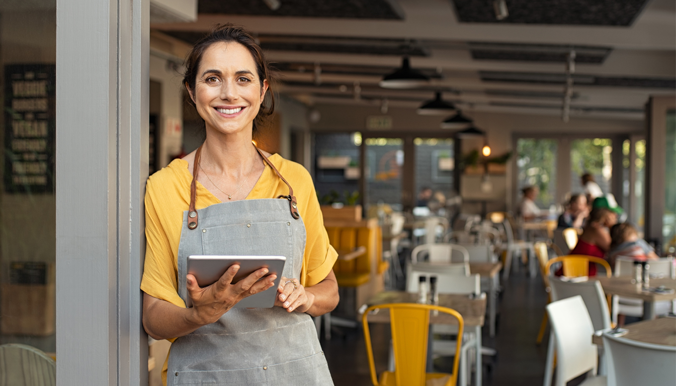 A smiling woman in an apron holds a tablet at the door of a busy cafe