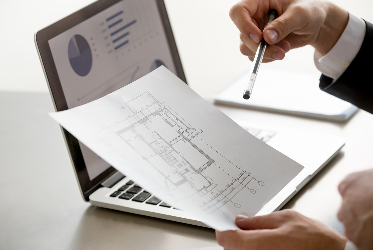 Hands holding a building floor plan and a pen in front of a laptop showing charts